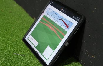 A tablet displaying a baseball hit trajectory graphic is propped against a black wall on artificial turf.