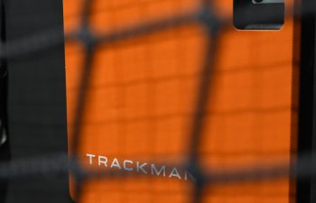 An orange TrackMan device with a visible sensor and indicator light is mounted indoors, partially obscured by a net in the foreground.