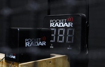 A Pocket Radar speed measurement device is set up on a wooden box, displaying a blank digital screen, with a black background and netting partially visible in the foreground.