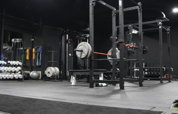 A gym with a power rack, barbell loaded with weights, dumbbells on racks, exercise balls, and colorful resistance bands hanging on the wall.