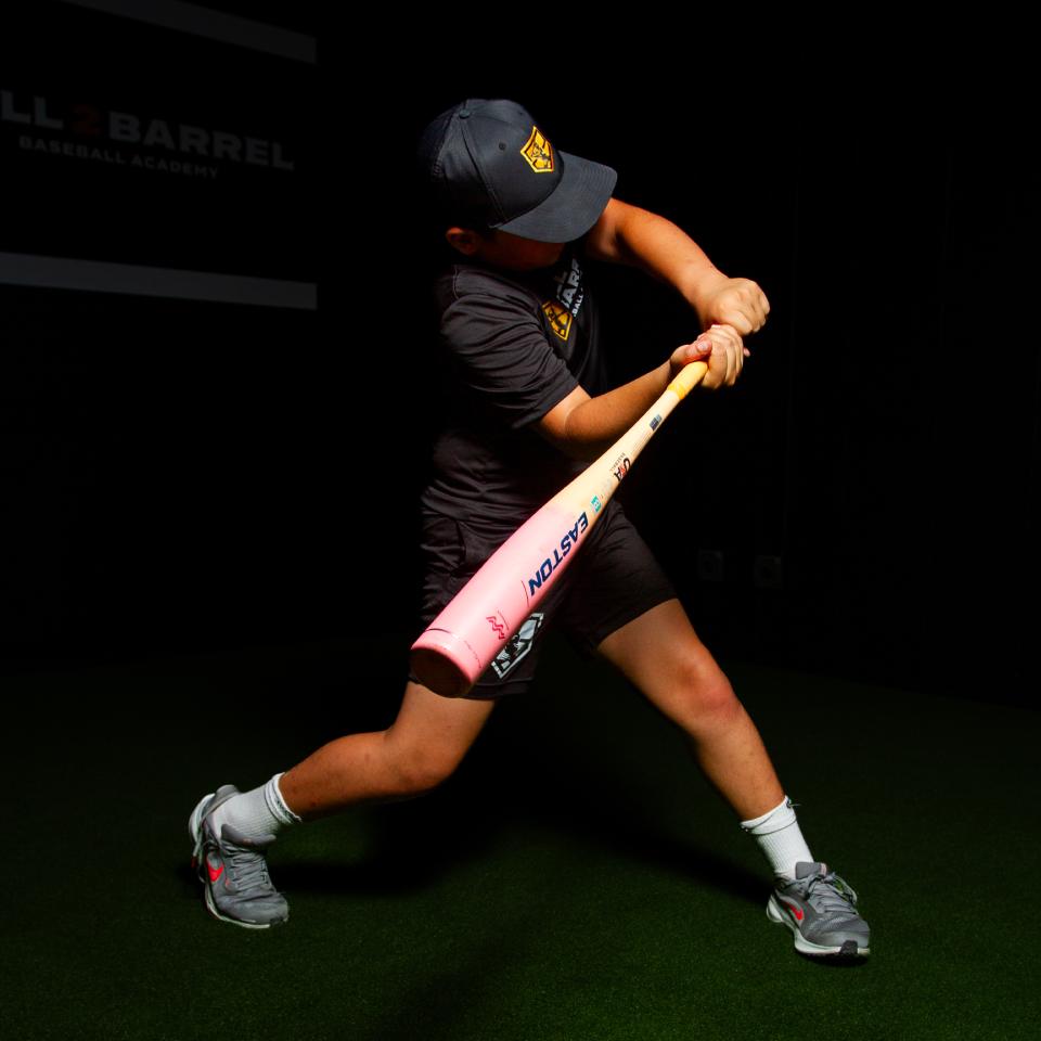 A young baseball player in a dark uniform and cap swings a pink Easton bat indoors on artificial turf.