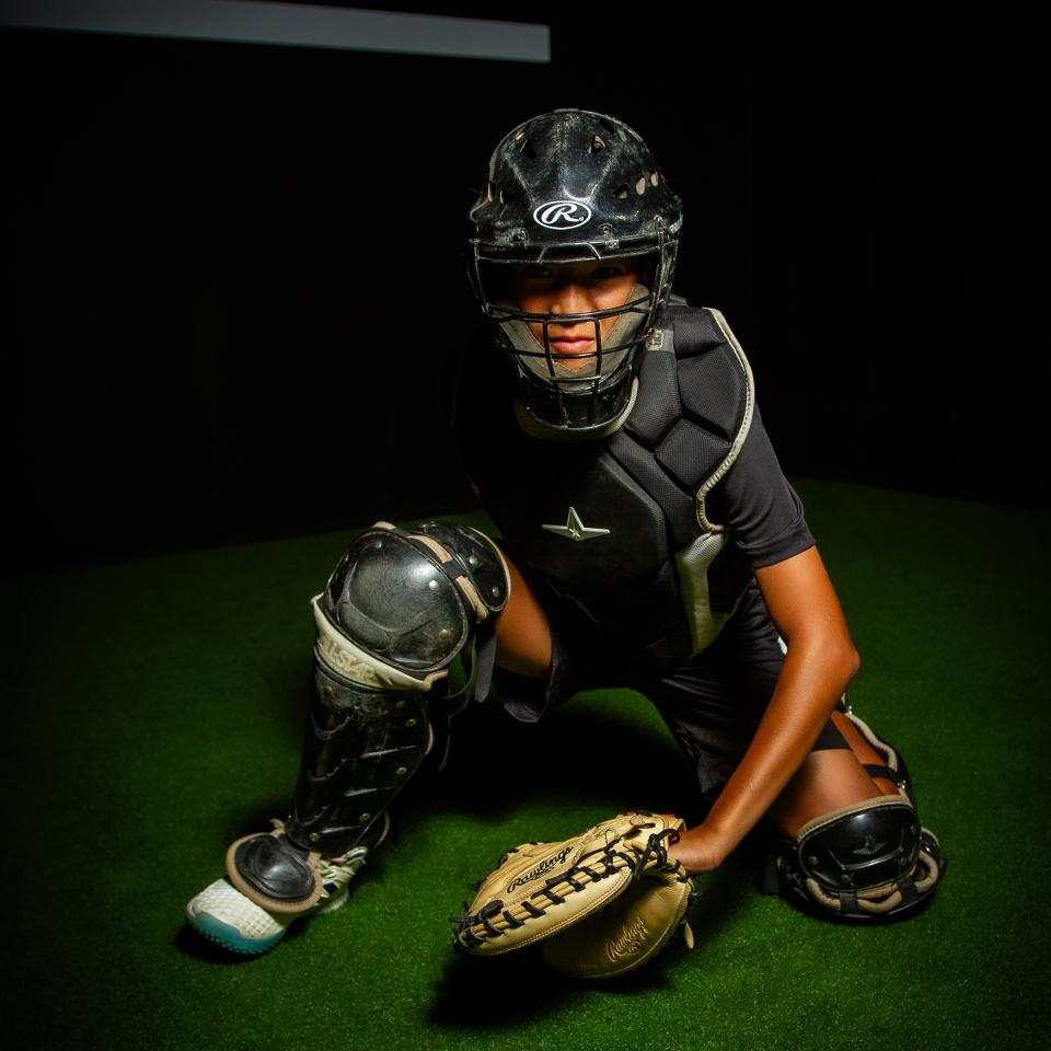 A baseball catcher in full protective gear crouches on artificial turf, holding a mitt and wearing a helmet and chest protector.