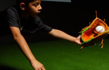 A boy dives on artificial turf, reaching out with an orange baseball glove to catch a baseball.