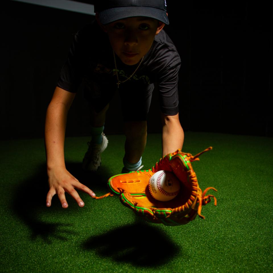 A young boy wearing a cap and dark shirt catches a baseball with a glove while leaning forward on artificial turf under dramatic lighting.