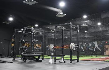 A gym with weightlifting equipment, including a bench press and squat rack, under bright overhead lights on a rubberized floor.